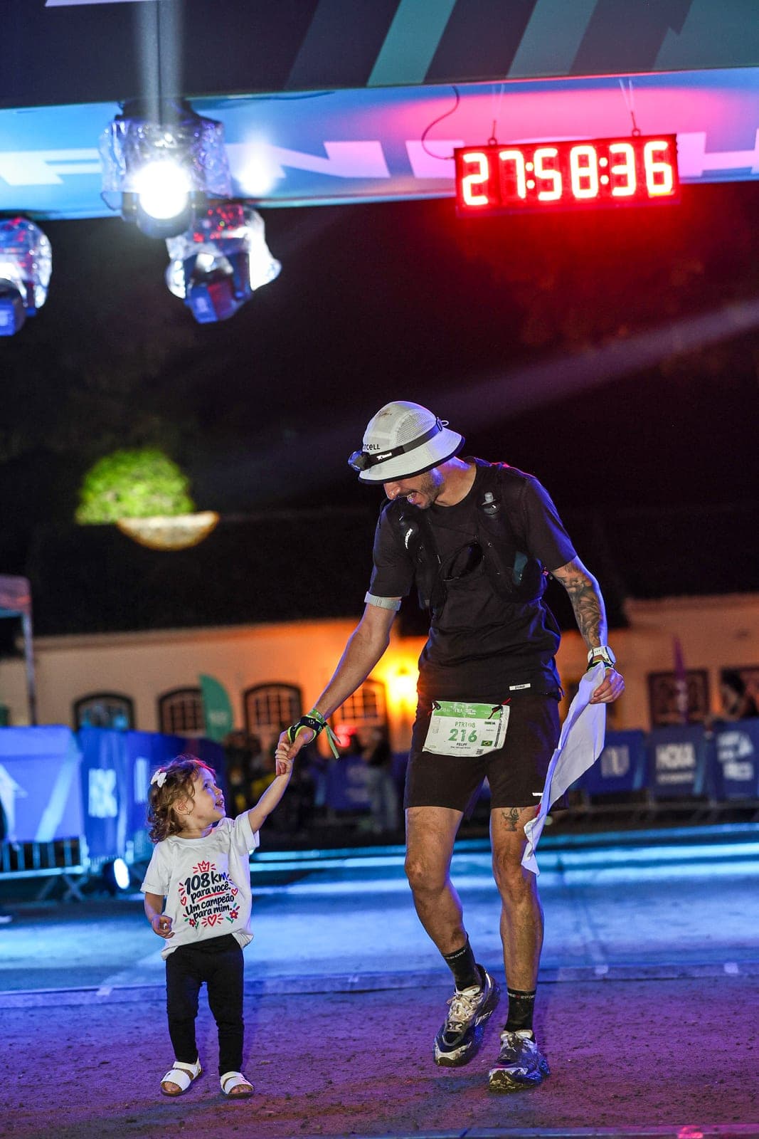 Felipe Bessa de mãos dadas com a filha pequena na chegada de uma prova noturna, sob as luzes do pórtico.